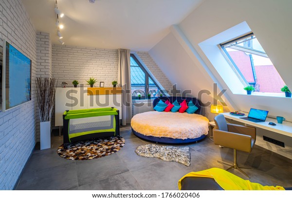 Modern loft interior of bedroom in apartment. Roof\
window. Round bed. Workplace. TV on the brick wall. Playpen. Home\
decor.