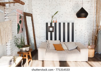 Modern loft interior with sofa, studio lamp, mirror, stripes on white brick wall and flowers in pot.