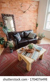 Modern Loft Living Room With High Ceiling. Leather Sofa, Wooden Table, Monstera, Book, Wicker Rug And Clay Dishes, Mirror With Light Bulbs, Wooden Floor, Deer Antlers On A Red Brick Wall, Red Carpet