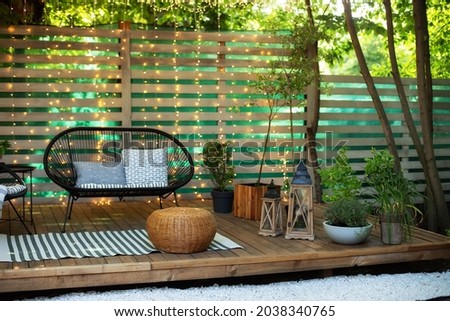 Modern lounge outdoors in backyard. Terrace house with plants, wooden wall and table, comfortable sofa, armchair and lanterns. Cozy space in patio or balcony. Wooden verande with garden furniture. 
