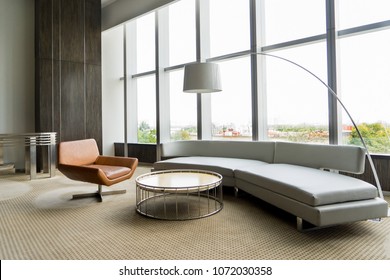 Modern Lounge Room Interior In Office Building. Room With Panoramic Window, Modern Leather Chair, White Sofa And Floor Lamp. Downtown Workspace Design Concept