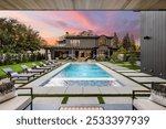 Modern luxury home with a swimming pool and sun loungers at sunset, featuring a well-manicured garden and outdoor seating area.