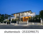 Modern luxury house exterior with large windows, illuminated at dusk, surrounded by greenery.