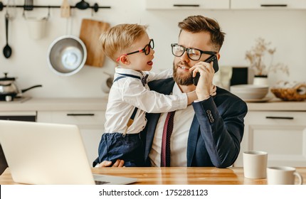 Modern male entrepreneur in formal outfit sitting at table with laptop and speaking on mobile phone while playful son asking for attention during remote work from home