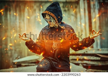 A modern male shaman in black ethnic cloak with dreadlocks, tattoos and sunglasses meditates on the street surrounded by sparks of fire. Shamanic ritual. 