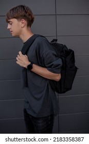 The Modern Male Teenager Dressed Black T-shirt, Jeans, Sunglasses, Watches And Bagback Walking In The Street At The Background Grey Wall
