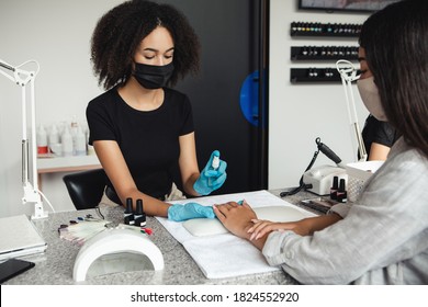 Modern Manicure And Health Protection In Beauty Salon During Epidemic. African American Woman In Protective Mask And Gloves Disinfects Hands Of Asian Client On Table With Nail Equipment, Free Space