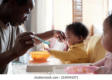 Modern mature African American man taking care of his twin baby daughters feeding them with delicious fruit puree - Shutterstock ID 2145941071
