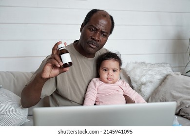 Modern Mature Black Man Sitting On Sofa In Front Of Laptop With Baby Daughter On His Lap Having Online Consultation With Pediatrician