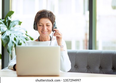 Modern Mature Woman Wearing, Holding Headset Sitting At Table In Front Of Computer, Laptop Looking At It, Smiling Happily. Positive Freelancer, Customer Representative Working Happy