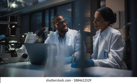 Modern Medical Research Laboratory: Caucasian and Black Scientists Work on Laptop, Do Data Analysis, Talk. Advanced Scientific Pharmaceutical Lab for Medicine, Biotechnology Development. Evening Time