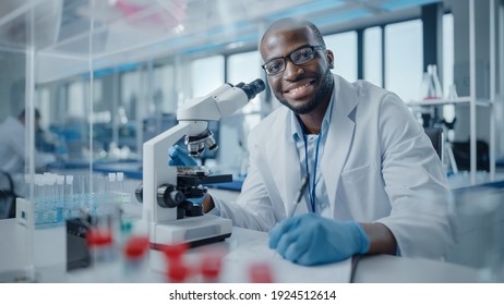 Modern Medical Research Laboratory: Portrait Of Male Scientist Using Microscope, Charmingly Smiling On Camera. Advanced Scientific Lab For Medicine, Biotechnology, Microbiology Development