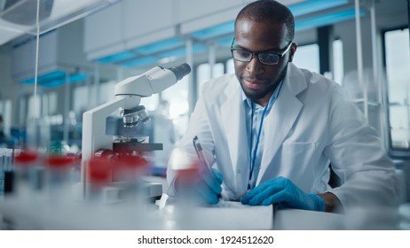 Modern Medical Research Laboratory: Portrait Of Male Scientist Using Microscope, Writing Down Analysis Information. Advanced Scientific Lab For Medicine, Biotechnology, Microbiology Development