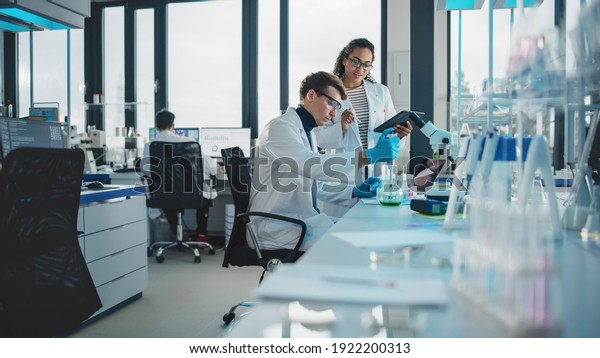 Modern Medical Research Laboratory: Shot of Two Young\
Scientists Using Pipette, Digital Tablet, Doing Sample Analysis,\
Talking. Diverse Team of Specialists work in Advanced Bio\
Technology Lab