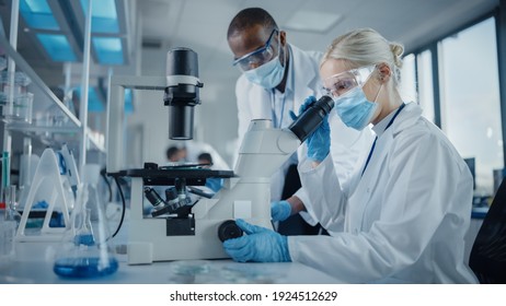 Modern Medical Research Laboratory: Two Scientists Wearing Face Masks Working Together Using Microscope, Analysing Samples, Talking. Advanced Scientific Lab for Medicine, Biotechnology.