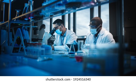 Modern Medical Research Laboratory: Two Scientists Wearing Face Masks use Microscope, Analyse Sample in Petri Dish, Talk. Advanced Scientific Lab for Medicine, Biotechnology. Blue Color