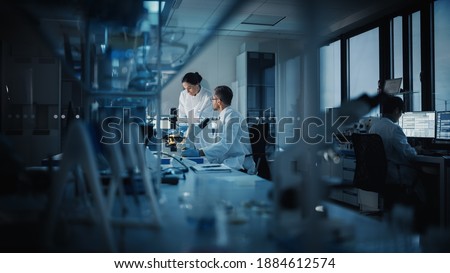 Modern Medical Research Laboratory: Two Scientists Working, Using Digital Tablet, Analyzing Test, Talking. Advanced Scientific Pharmaceutical Lab for Medicine, Biotechnology Development. Evening Time