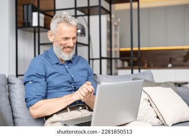 Modern middle-aged bearded man using laptop computer sitting on the couch at home, spending leisure time online, watching movies. Mature male working remotely from home, looks at the screen and smiles - Powered by Shutterstock