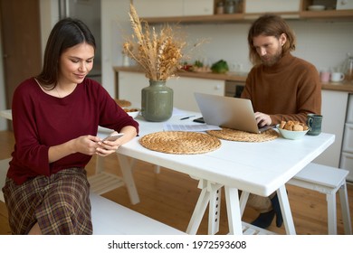 Modern Millenials Using Electronic Devices. Busy Young Bearded Manager Working On Laptop Sitting At Table With His Joyful Wife Smm Expert Who Is Typing New Post For Social Media Account On Cell Phone