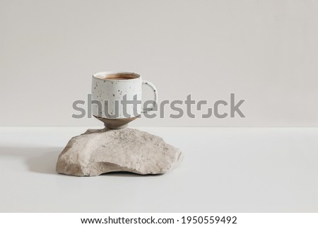 Modern minimal lifestyle still life scene. Speckled ceramic cup of coffee standing on sandstone rock, white table. Beige wall background. Breakfast drink concept. Empty copy space, no people.