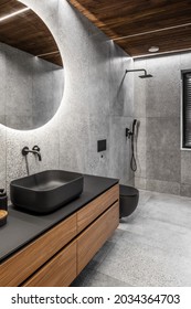 Modern Minimalist Bathroom Interior With Stone Grey Tiles, Wooden Furniture, Black Sink And Toilet And Round Mirror. Spa Bathroom Concept