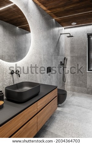 Modern minimalist bathroom interior with stone grey tiles, wooden furniture, black sink and toilet and round mirror. Spa bathroom concept