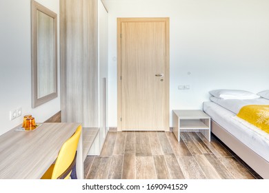 Modern Minimalist Hotel Room With Closed Bathroom Door, Mirror, Glasses, Yellow Chair And Wardrobe. Wooden Tile Floor. Plain White Wall Without Decoration.