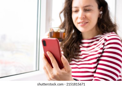 Modern Mobile Phone In Red Case Close Up In Hand Indoor. Smiling Brunette Woman Listening Music, Podcast, Watching Video, Making Video Call, Browsing Website, Checking Email, Drinking Tea. Copy Space