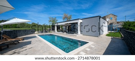 Modern modular house backyard featuring a rectangular swimming pool, large patio with wooden sun loungers, and umbrellas against a blue sky. Contemporary prefabricated home design