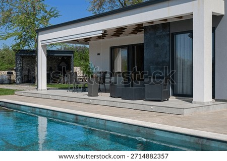 Modern modular house backyard featuring a rectangular swimming pool, large patio with wooden sun loungers, and umbrellas against a blue sky. Contemporary prefabricated home design