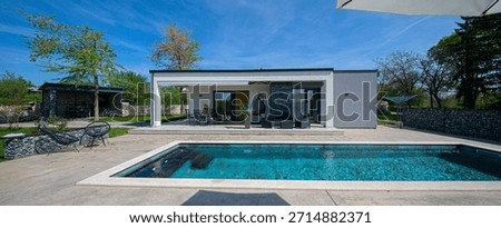 Modern modular house backyard featuring a rectangular swimming pool, large patio with wooden sun loungers, and umbrellas against a blue sky. Contemporary prefabricated home design