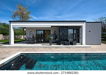 Modern modular house backyard featuring a rectangular swimming pool, large patio with wooden sun loungers, and umbrellas against a blue sky. Contemporary prefabricated home design
