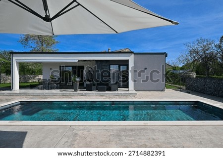 Modern modular house backyard featuring a rectangular swimming pool, large patio with wooden sun loungers, and umbrellas against a blue sky. Contemporary prefabricated home design