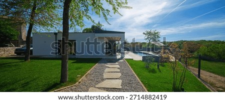 Modern modular house backyard featuring a rectangular swimming pool, large patio with wooden sun loungers, and umbrellas against a blue sky. Contemporary prefabricated home design
