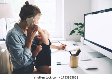 Modern Mom Balances Between Work And Child Home Schooling On Sick Leave Or Quarantine. Mother Works From Home With Kid. Woman Responding On Phone Calls, Working On Computer With A Toddler On Her Lap.