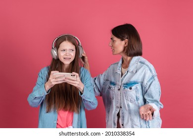 Modern Mom And Daughter In Denim Jackets On Terracotta Background Girl In Headphones Play Game On Phone And Disappointed Angry Mom Stands By