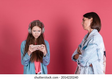 Modern Mom And Daughter In Denim Jackets On Terracotta Background Girl In Headphones Play Game On Phone And Disappointed Angry Mom Stands By