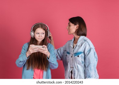 Modern mom and daughter in denim jackets on terracotta background girl in headphones play game on phone and disappointed angry mom stands by