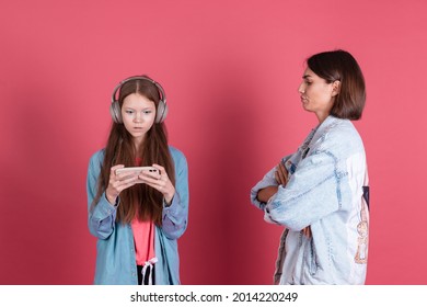 Modern Mom And Daughter In Denim Jackets On Terracotta Background Girl In Headphones Play Game On Phone And Disappointed Angry Mom Stands By