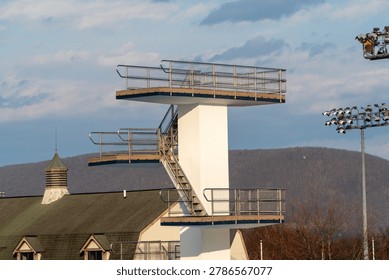 Modern multi-level diving platform with stainless steel railings and wooden platforms at an outdoor aquatic sports complex, illuminated by golden hour light against a backdrop of forested mountains and a partly cloudy sky, symbolizing sports infrastructure and readiness.