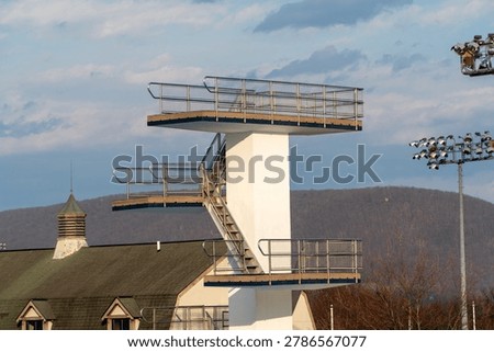 Modern multi-level diving platform with stainless steel railings and wooden platforms at an outdoor aquatic sports complex, illuminated by golden hour light against a backdrop of forested mountains and a partly cloudy sky, symbolizing sports infrastructure and readiness.