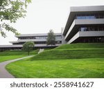 A modern multi-story concrete building with a geometric design and grid windows, set against a lush lawn with two small trees. A winding path leads to the entrance, framed by soft, overcast light.