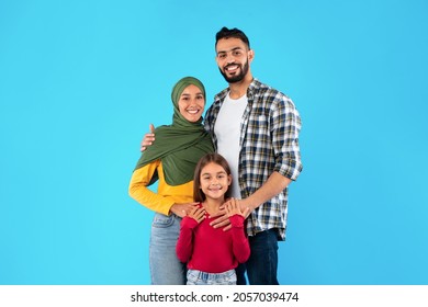 Modern Muslim Family Of Three Hugging Smiling To Camera Standing Together Over Blue Studio Background. Islamic Mother In Hijab And Father Embracing Their Happy Little Daughter