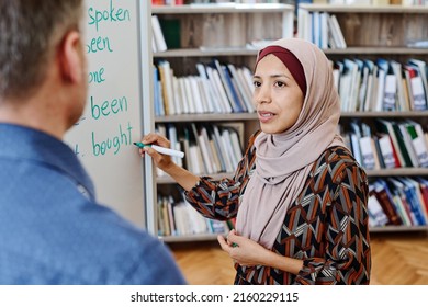 Modern Muslim woman wearing hijab standing at whiteboard doing task with irregular verbs and answering teachers question during English lesson - Shutterstock ID 2160229115