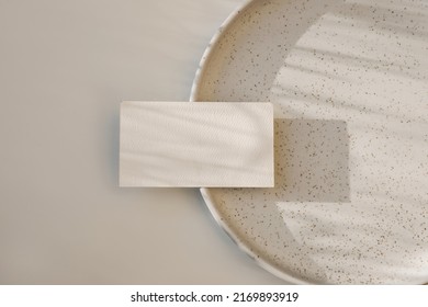 Modern neutral branding stationery. Closeup of textured blank business card, invitation mockup on dotted, speckled ceramic plate in sunlight. Beige table background. Flat lay, top view. No people.