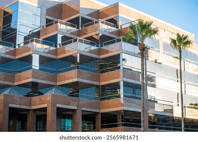 Modern office building with angular architecture, reflective glass windows, brown stone facade, and palm trees under bright daylight.