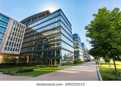 Modern office buildings with large glass windows reflecting sunlight. A well-maintained pathway runs alongside green lawns and trees, creating a blend of architecture and nature. - Powered by Shutterstock