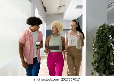 In modern office, diverse young team discussing while walking. African American man holding coffee, biracial woman carrying tablet, Caucasian woman with thoughtful expression on blue background, - Powered by Shutterstock