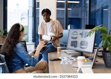 In modern office, diverse young team exploring virtual reality. African American male manager showing VR headset to Caucasian female coworker, both surrounded by tech gadgets, unaltered - Powered by Shutterstock