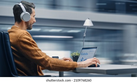 Modern Office: Handsome Businessman Sitting At His Desk Working On A Laptop Computer. Man Wearing Headphones, Listens To Music, Podcast. Motion Blur Background Showing Active Work Day.